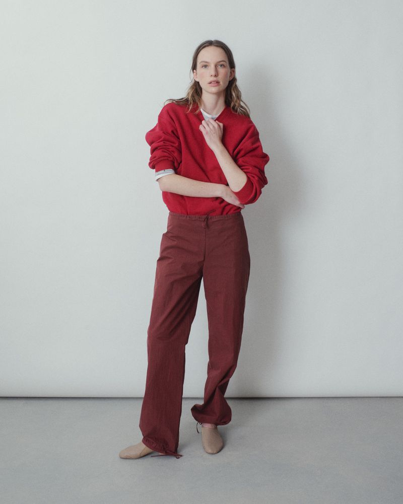 A woman in a red sweatshirt and maroon pants stands in front of a white wall in a studio, looking directly at the camera.