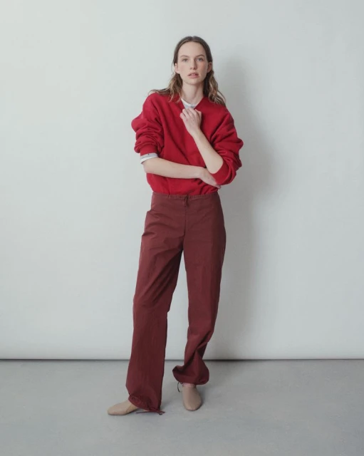 A woman in a red sweatshirt and maroon pants stands in front of a white wall in a studio, looking directly at the camera.