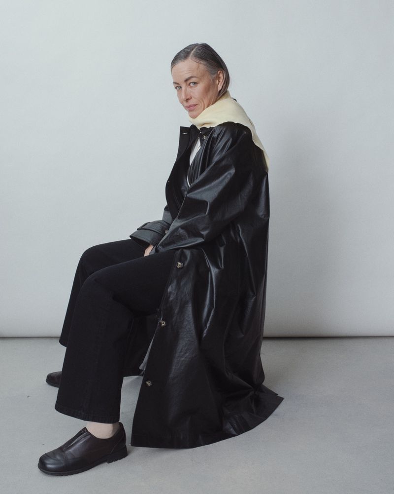 Stylish mature woman with gray hair and a black coat seated against a neutral background in a fashion forward portrait.