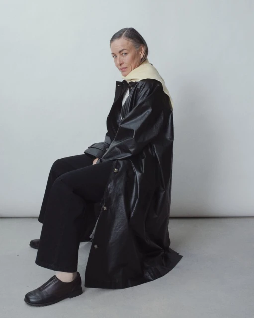 Stylish mature woman with gray hair and a black coat seated against a neutral background in a fashion forward portrait.