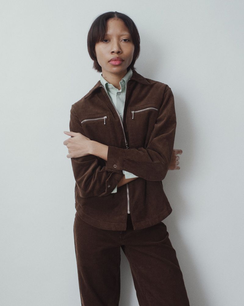 A young adult posing in a brown corduroy jacket and pants in a studio. The subject faces forward with arms crossed and a neutral expression.