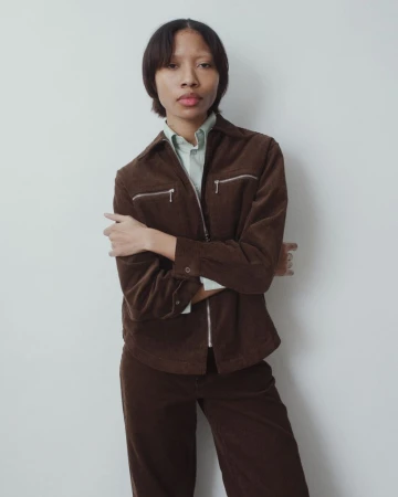 A young adult posing in a brown corduroy jacket and pants in a studio. The subject faces forward with arms crossed and a neutral expression.