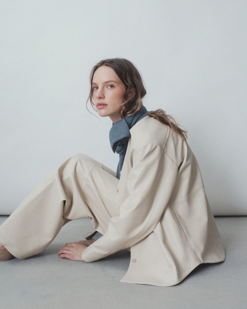 Young woman with long wavy hair sits indoors wearing a cream-colored jacket and matching pants with a blue scarf, posing against a plain white background.