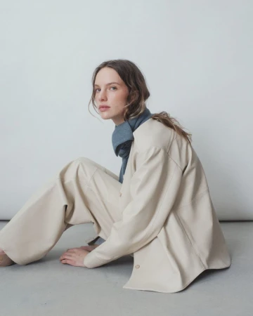 Young woman with long wavy hair sits indoors wearing a cream-colored jacket and matching pants with a blue scarf, posing against a plain white background.