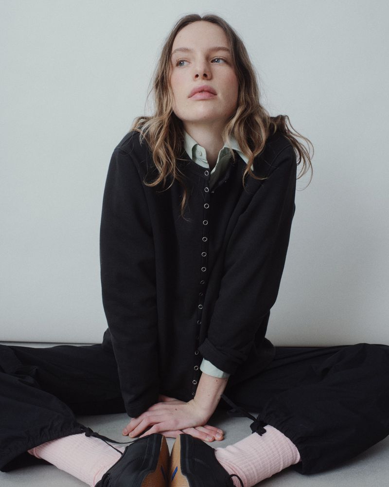 A woman with long, wavy hair sits cross-legged in a studio wearing a black button-down cardigan, light green shirt, and black pants. She wears pink socks.