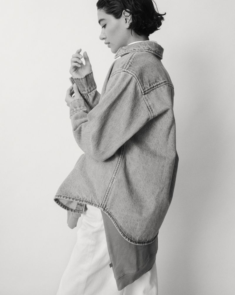 A woman in a light denim jacket poses against a white wall in a monochrome shot. The jacket is oversized, sleeves rolled up.