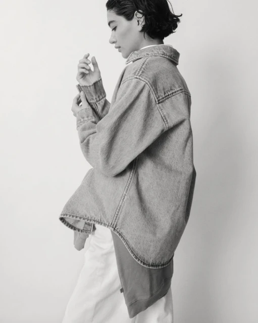 A woman in a light denim jacket poses against a white wall in a monochrome shot. The jacket is oversized, sleeves rolled up.