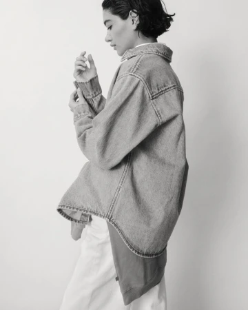 A woman in a light denim jacket poses against a white wall in a monochrome shot. The jacket is oversized, sleeves rolled up.