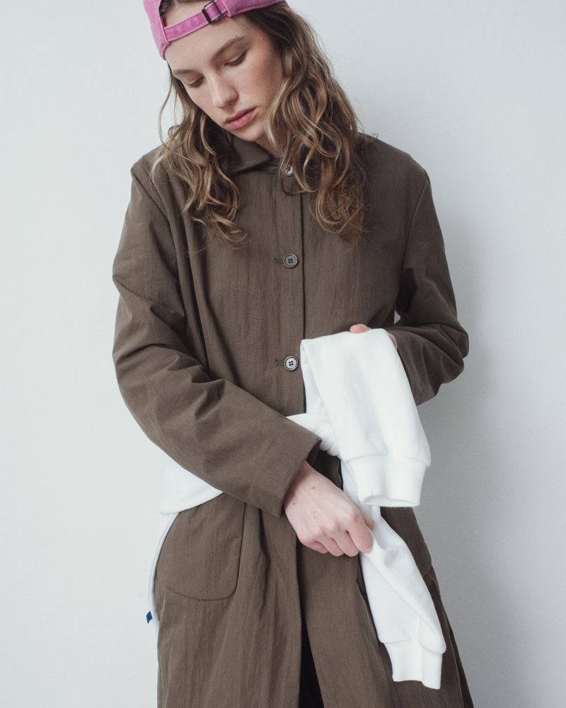 Adult woman poses indoors wearing a brown coat and pink baseball cap, holding a white long-sleeve shirt against a white background.