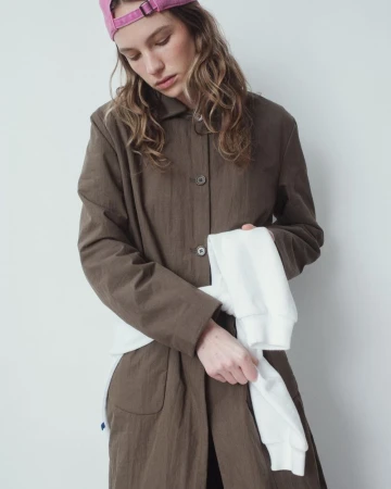 Adult woman poses indoors wearing a brown coat and pink baseball cap, holding a white long-sleeve shirt against a white background.