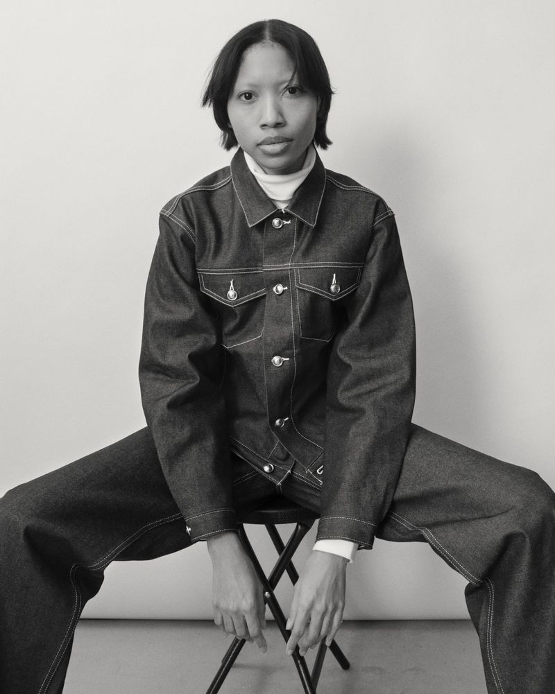 Monochrome portrait of a Black woman in a denim jacket and pants, sitting on a stool against a white background, fashion and portrait photography.