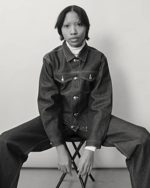 Monochrome portrait of a Black woman in a denim jacket and pants, sitting on a stool against a white background, fashion and portrait photography.