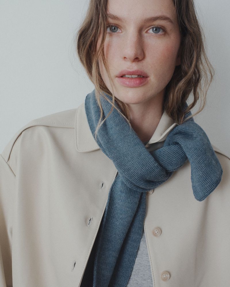 Close-up of a fair-skinned woman wearing a blue knitted scarf and a beige trench coat in a bright indoor fashion shot.