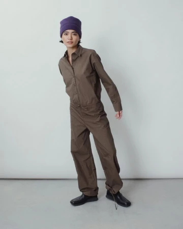 Model wearing a purple beanie hat and olive brown buttoned jumpsuit with cinched hems against a plain light studio backdrop.