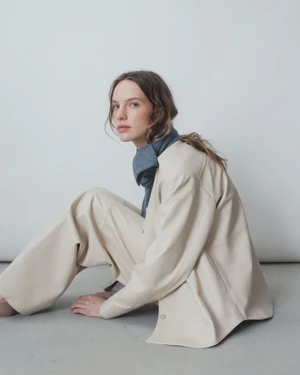 Model seated on the floor wearing an oversized cream jacket, matching wide-leg trousers, and a blue neck scarf against a neutral studio backdrop.
