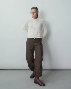 Model wearing eyeglasses, an off-white zip-up knit hoodie, brown loose-fitting trousers, and brown ankle boots in a studio setting.