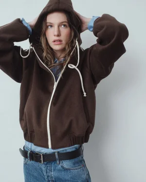 Model wearing a brown hooded zip-up jacket over a blue collared shirt and denim jeans against a neutral studio backdrop.