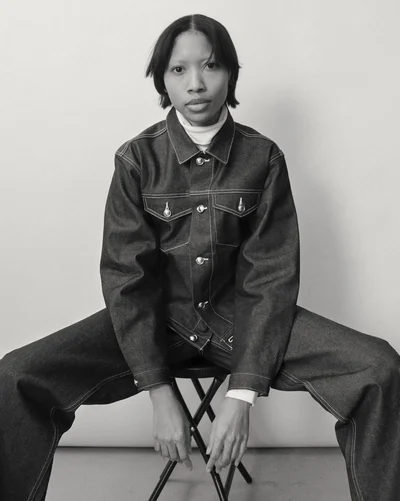 Model Dominique Babineaux seated on a stool wearing a dark denim jacket with contrast stitching and matching wide-leg trousers.