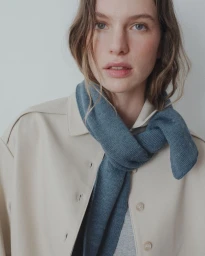 Model wearing a knotted slate blue knit scarf over a beige collared jacket against a neutral studio background.