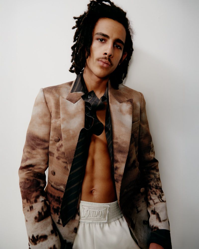 Portrait of a young adult male in a printed suit jacket, unbuttoned dark shirt, and white shorts with 'LONDON' text, against a white wall.