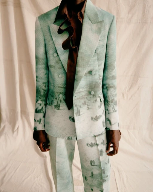 Fashionable male model poses in a light green suit with a nature scene print and ruffled brown shirt against a neutral backdrop.