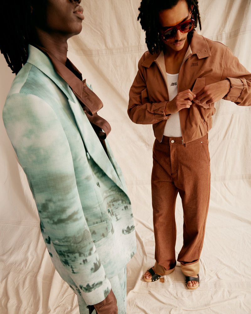 Two adult men in stylish outfits pose indoors against a neutral backdrop. One wears a printed suit, the other, a jacket and trousers.
