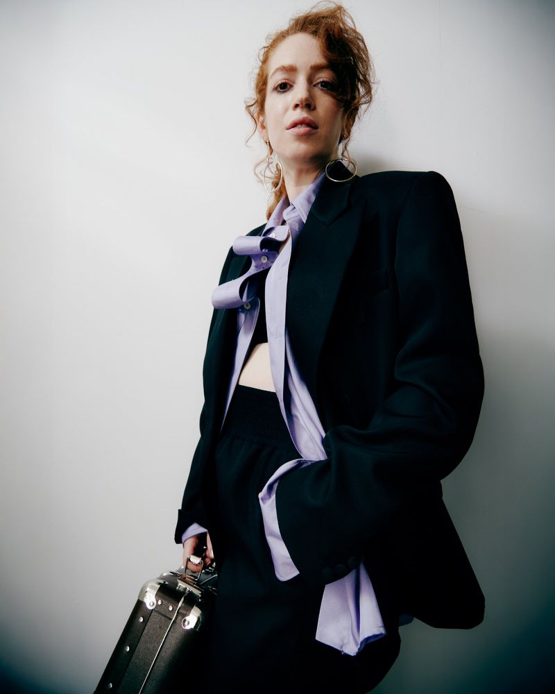 Adult woman with red curly hair poses in a black blazer and skirt, holding a briefcase, against a plain white background in studio lighting.