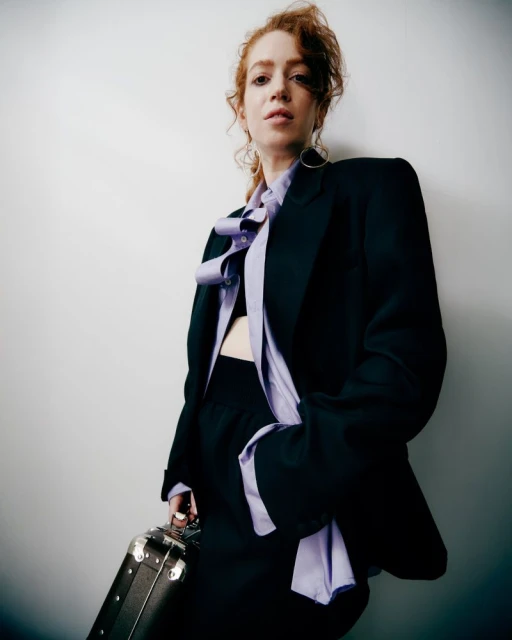 Adult woman with red curly hair poses in a black blazer and skirt, holding a briefcase, against a plain white background in studio lighting.