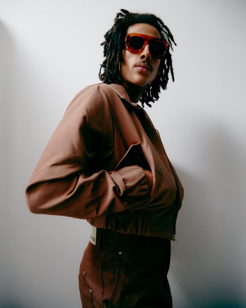 Portrait of a young adult male with dreadlocks wearing a brown jacket and sunglasses, standing against a white wall in soft, natural light.