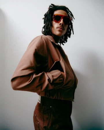 Portrait of a young adult male with dreadlocks wearing a brown jacket and sunglasses, standing against a white wall in soft, natural light.