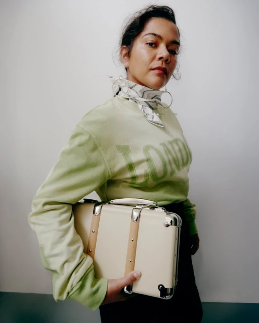Woman in a pistachio sweatshirt with the word 'London' holding a small vintage suitcase, posing in front of a plain wall.