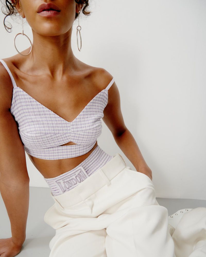 Woman with tan skin poses in a bralette and trousers indoors, featuring minimal makeup and gold earrings against a neutral background.