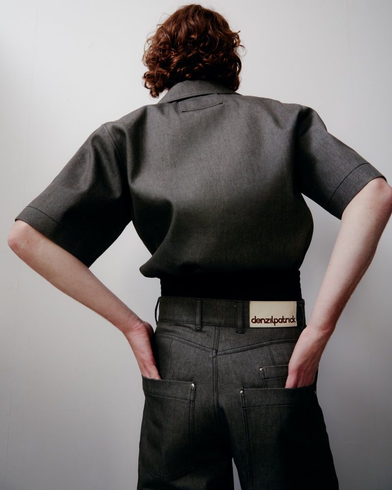 Back view of a person with red hair wearing a gray short-sleeved shirt and gray jeans against a plain white background in a studio shot.