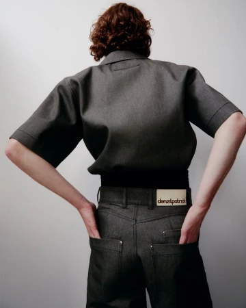 Back view of a person with red hair wearing a gray short-sleeved shirt and gray jeans against a plain white background in a studio shot.