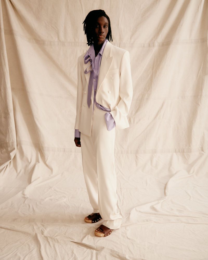 Fashionable adult male in cream suit and lilac shirt strikes a confident pose against a neutral backdrop, showcasing high-end editorial style.