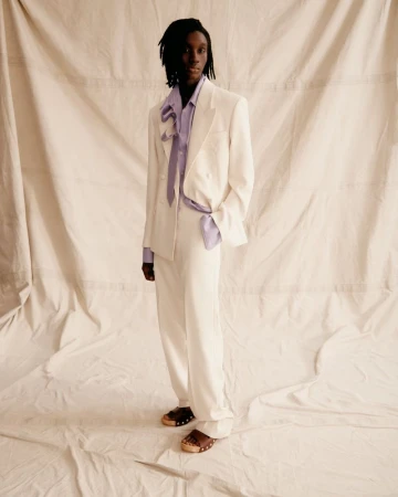 Fashionable adult male in cream suit and lilac shirt strikes a confident pose against a neutral backdrop, showcasing high-end editorial style.