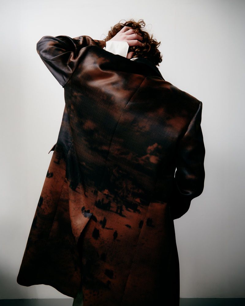 Back view of an adult with curly brown hair wearing a patterned brown coat with a white collared shirt against a light background.