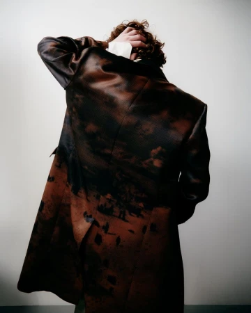Back view of an adult with curly brown hair wearing a patterned brown coat with a white collared shirt against a light background.