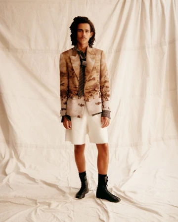Model wearing a landscape-printed tailored blazer, white shorts, and black lace-up boots against a draped fabric backdrop.