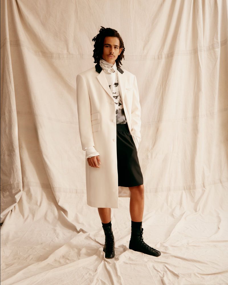 A male model wearing a long cream coat, black shorts, and black lace-up boots stands against a draped fabric backdrop.