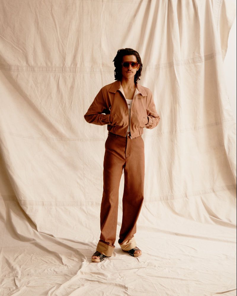 A stylish adult in sunglasses, a jacket, and brown pants poses against a neutral backdrop in a full shot, showcasing fashion and style.