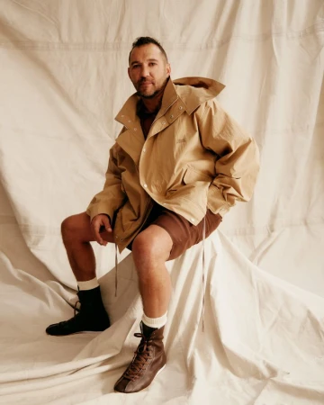 Adult male seated in a tan hooded jacket and brown shorts, wearing socks and lace-up boots, posing in front of a light-colored backdrop.