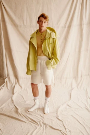 Male model in lime jacket and shorts poses in a studio against a cream-colored backdrop, showcasing modern casual fashion.