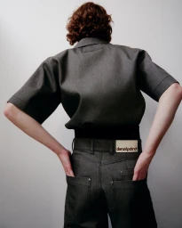 Model seen from behind wearing a short-sleeve grey shirt tucked into matching trousers with hands in back pockets.