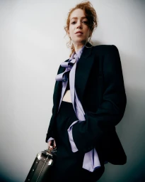 Model with red hair posing in an oversized black blazer, lavender shirt, and holding a black briefcase against a neutral background.