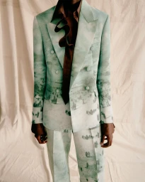 Model wearing a light green landscape print double-breasted suit with a brown ruffled necktie against a draped textile backdrop.