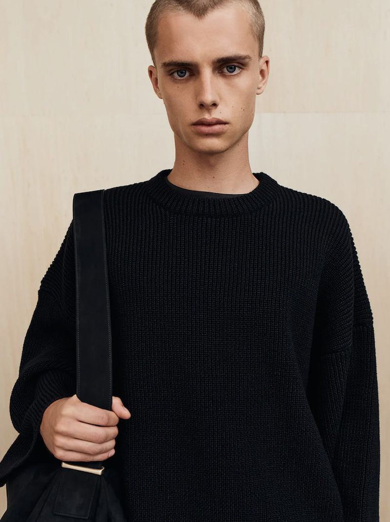Portrait of a young adult man wearing a black knit sweater and carrying a bag with a strap, standing indoors with neutral wall background.