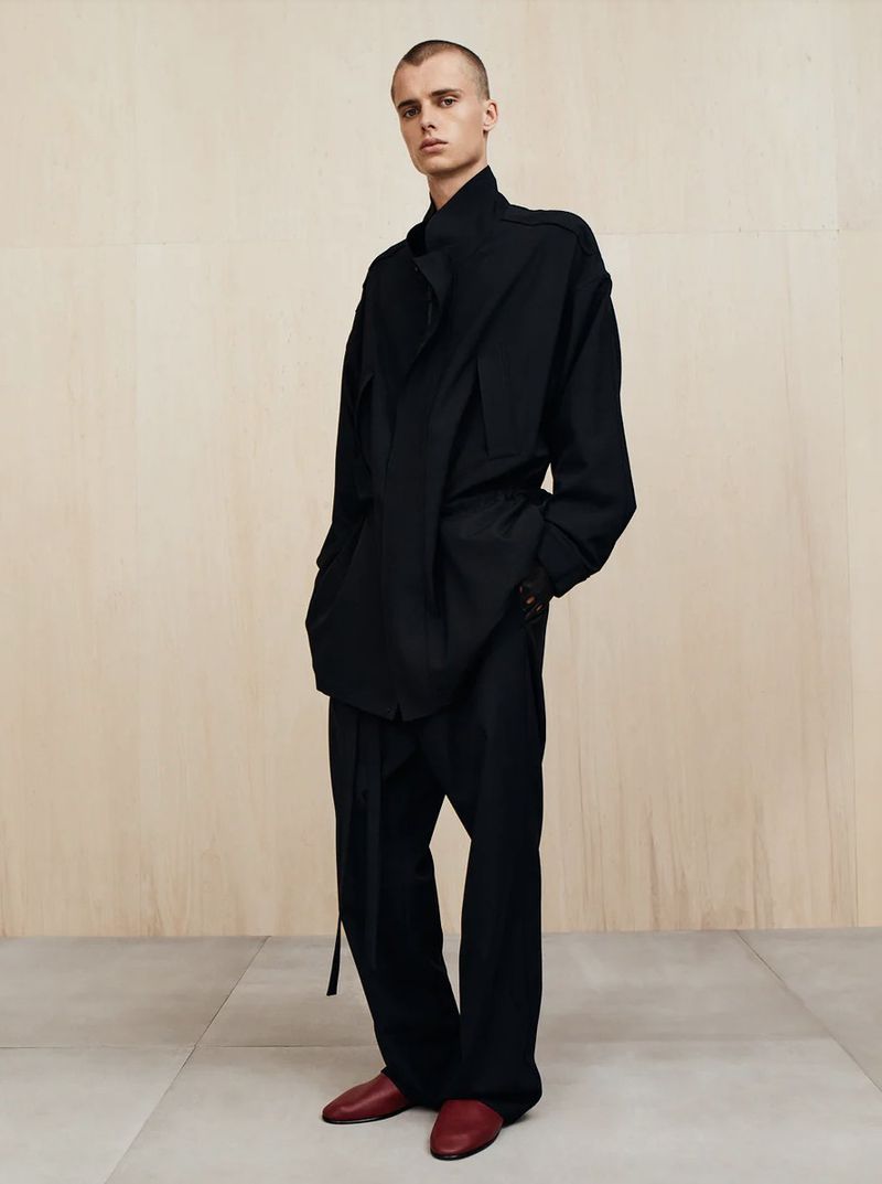 A single adult male stands in a studio wearing a black asymmetrical coat, black pants, and red shoes, with a neutral background.