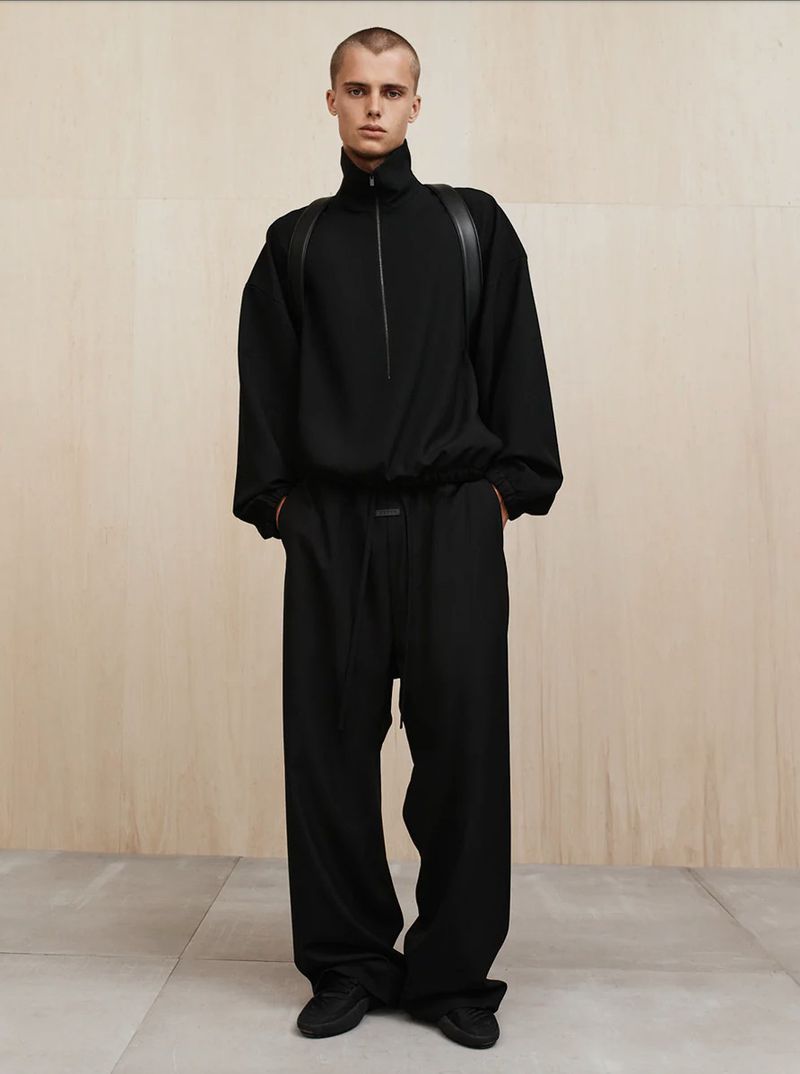 A slender person with a buzzcut is standing indoors, modeling a black long-sleeved shirt and wide-leg pants in front of a wood wall.
