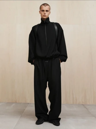 A slender person with a buzzcut is standing indoors, modeling a black long-sleeved shirt and wide-leg pants in front of a wood wall.